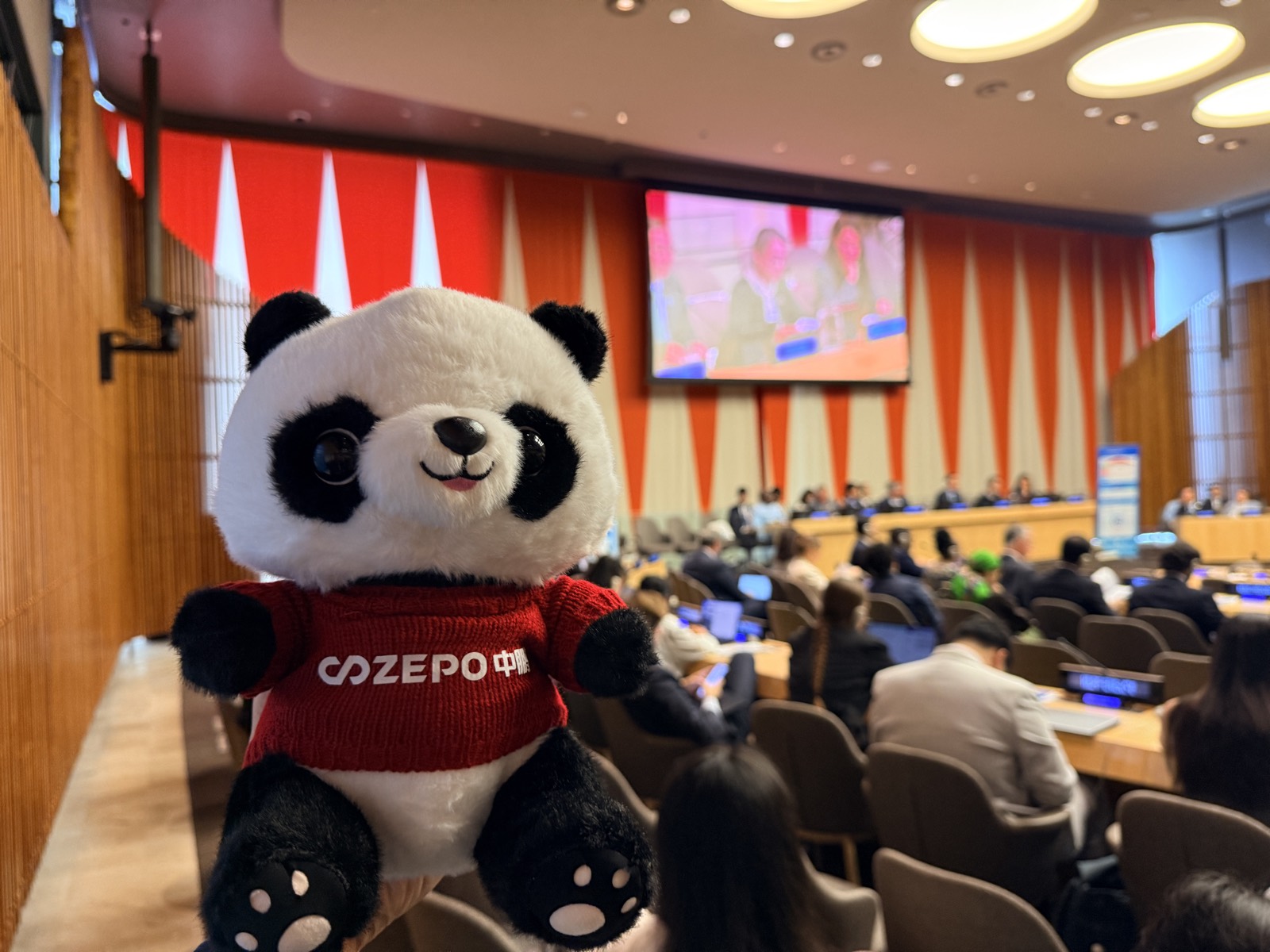 A plush panda mascot held up in the foreground of a UN conference room, with delegates seated at desks and a large screen showing the panel behind it.