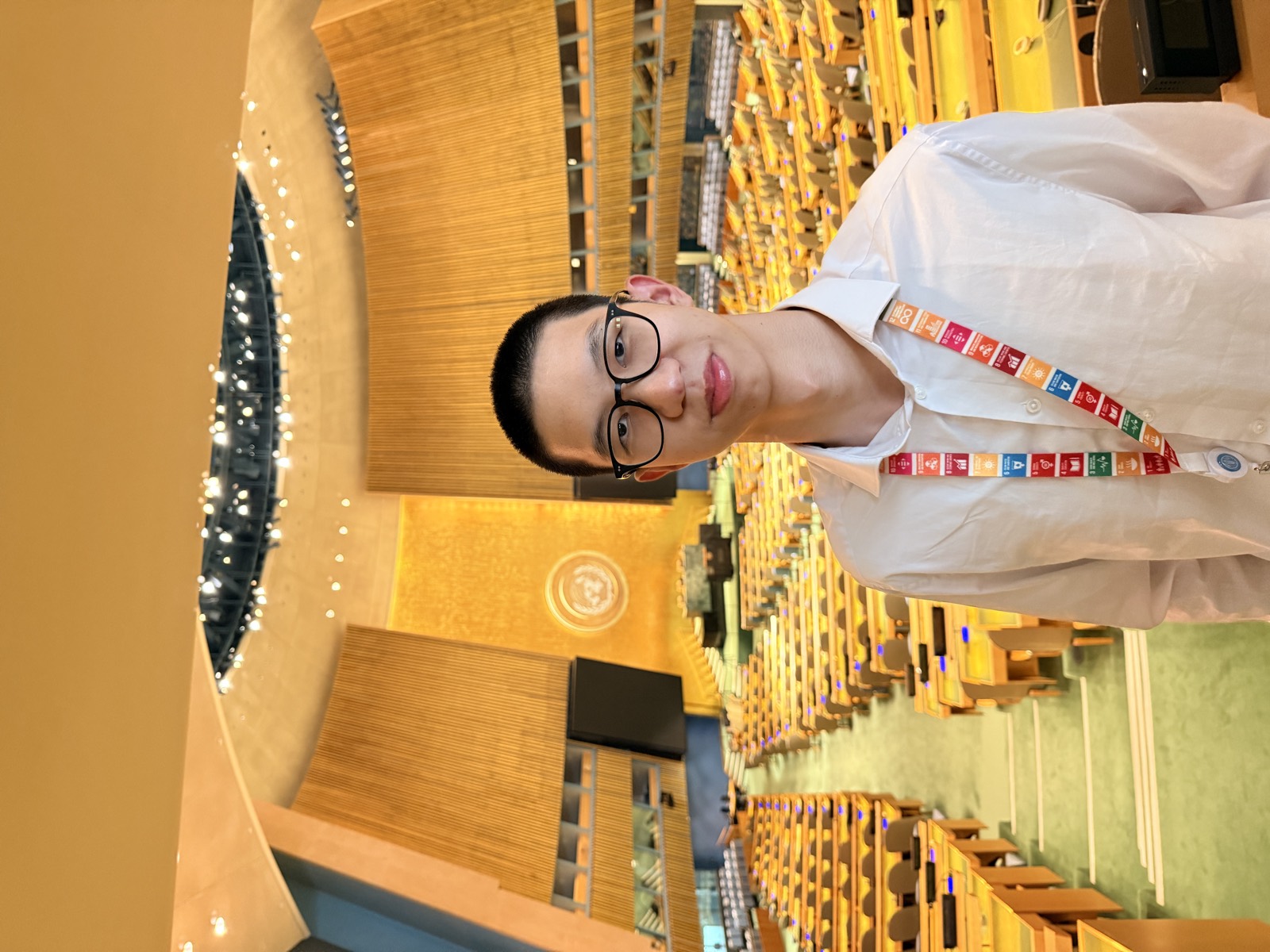 Yian Xie standing in the United Nations General Assembly Hall, wearing a lanyard printed with the Sustainable Development Goals, with rows of empty desks and the UN emblem behind him.